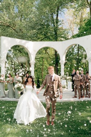 A bride and groom walk through an arch as guests toss flower petals at their outdoor wedding ceremony.