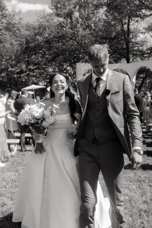 Black and white photo of smiling bride holding bouquet walking with groom after outdoor wedding ceremony.