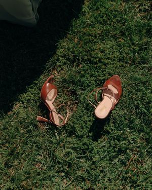 Brown leather heeled sandals with ankle straps resting on lush green grass in natural sunlight.
