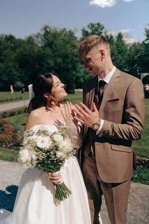 Newlywed couple showing wedding rings outdoors, bride holding white bouquet in elegant gown, groom in tan suit.