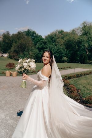 Radiant bride in off-shoulder gown twirls with bouquet in sunlit garden, veil flowing behind her.