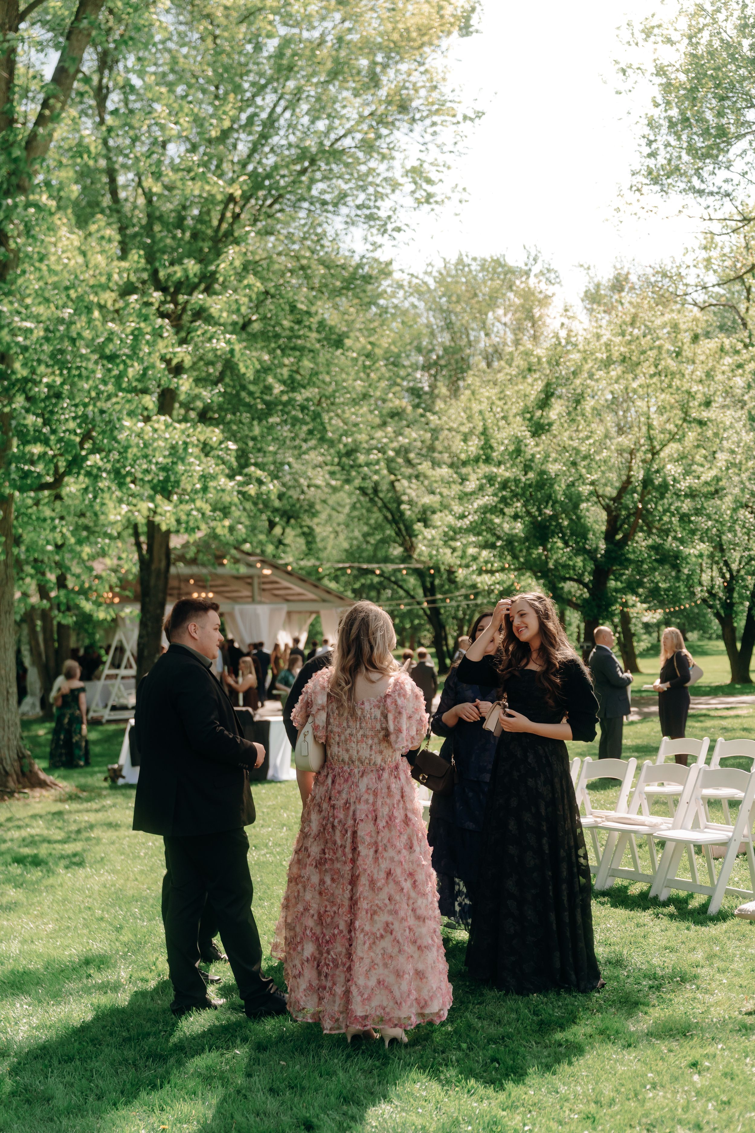 Guests in formal attire mingle on green lawn at outdoor wedding ceremony with white chairs and trees.
