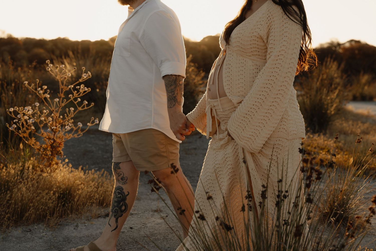 Expectant couple strolls through grassy location during a warm sunset maternity session at Rockingham Salt Lakes.