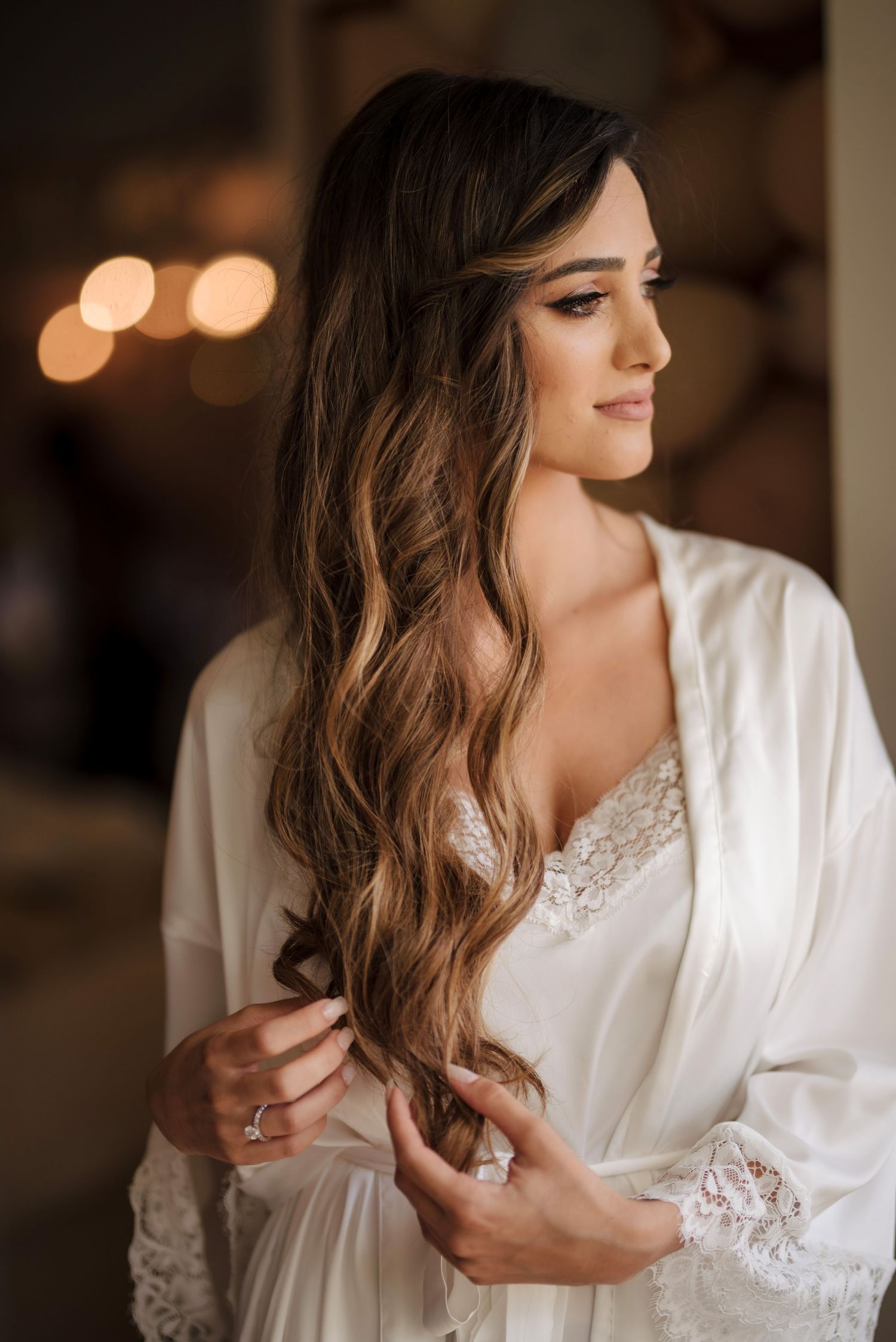 Bride in white lace robe with long wavy brunette hair, elegant bridal makeup, holding hair while looking away softly.