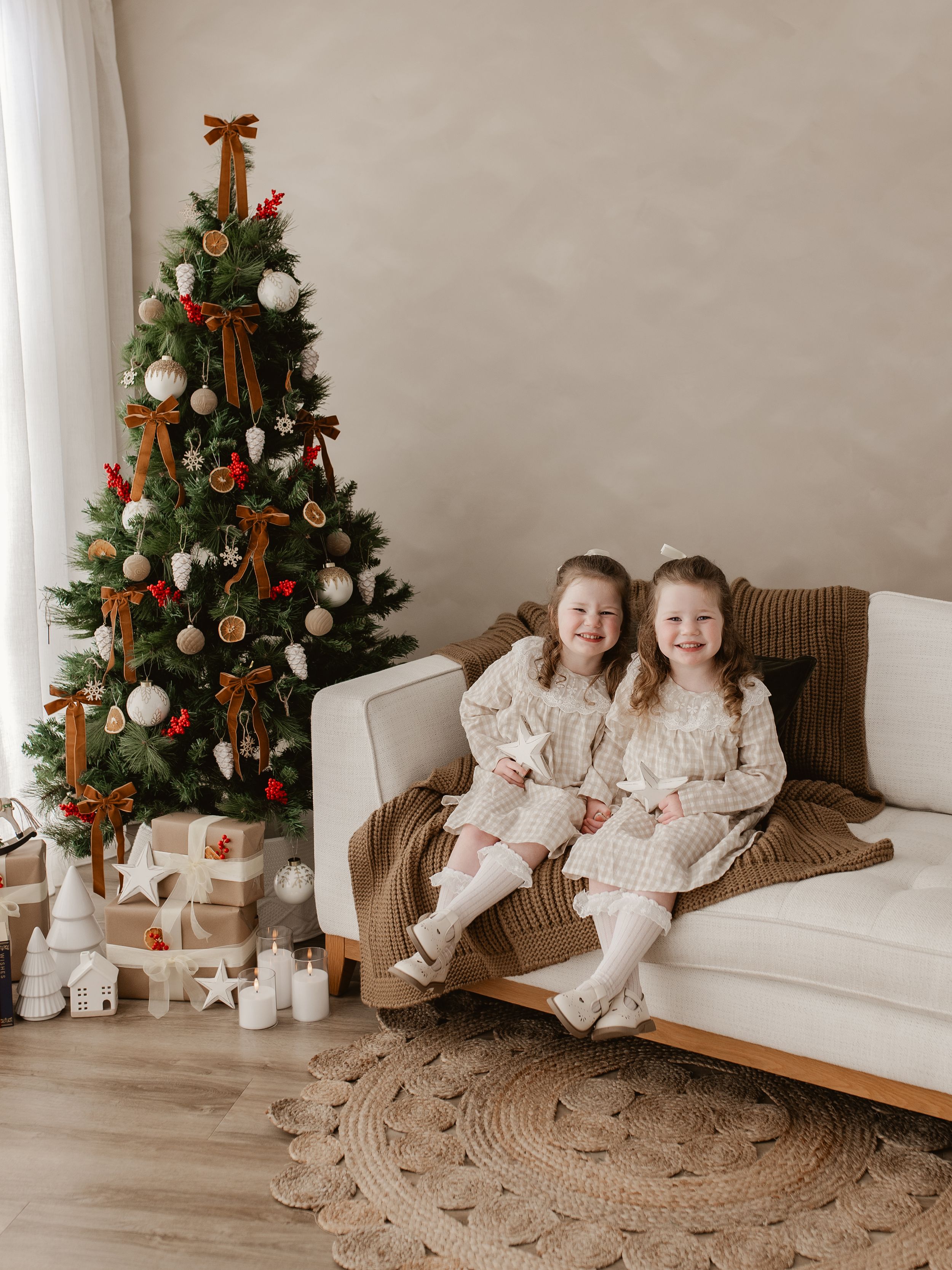 Two young girls in matching dresses smile on a white sofa beside a decorated Christmas tree with gifts.