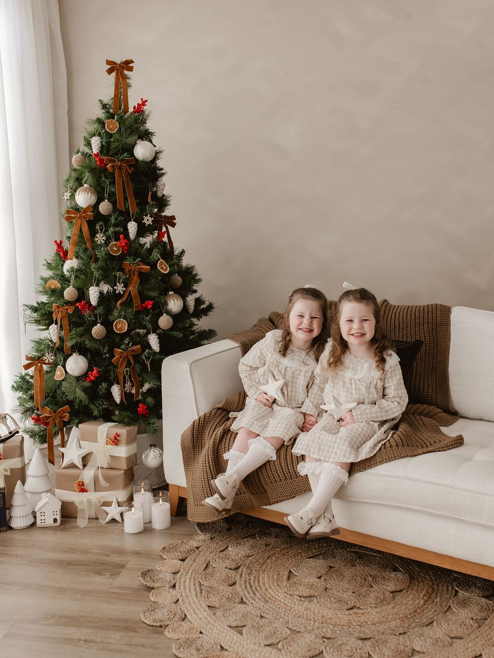 Two young girls in matching dresses smile on a white sofa beside a decorated Christmas tree with gifts.
