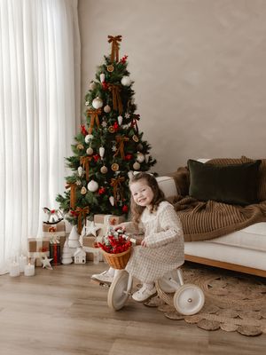 Young girl smiling on a toy tricycle beside a decorated Christmas tree with gifts in a cozy living room.