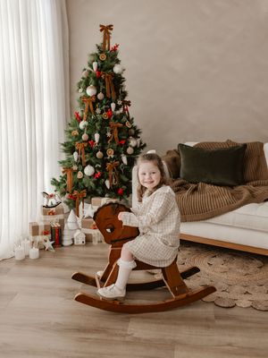 Young girl smiling on a wooden rocking horse beside a decorated Christmas tree with gifts in a cozy living room.