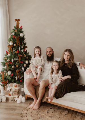Family of four posing by decorated Christmas tree on white couch, father with beard, two young daughters, festive holiday setting.