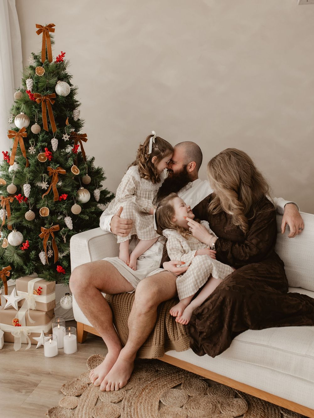 Family of four sharing a tender Christmas moment on a white sofa beside a decorated holiday tree with gifts.