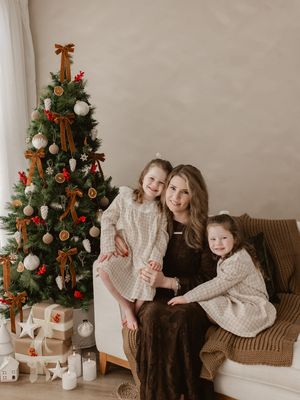 A mother and two daughters pose by a decorated Christmas tree with gifts in a cozy white living room.