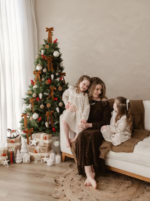 A mother and two daughters in cozy outfits sit by a decorated Christmas tree with gifts in a warm living room.