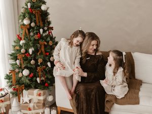 A mother and two daughters in cozy sweaters share a warm moment on a white sofa beside a decorated Christmas tree.