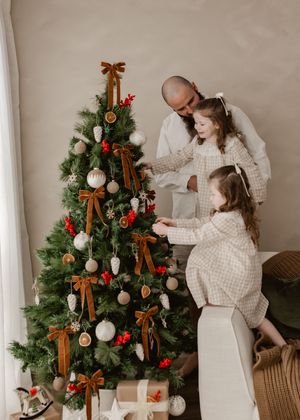A bald father and two young daughters in matching plaid dresses decorate a Christmas tree with bows and ornaments.