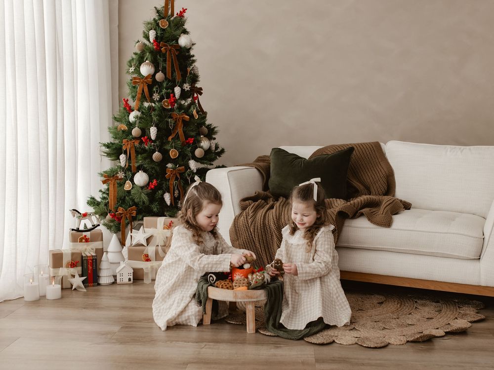 Two young girls in white dresses play with toys near a decorated Christmas tree and gifts in a cozy living room.