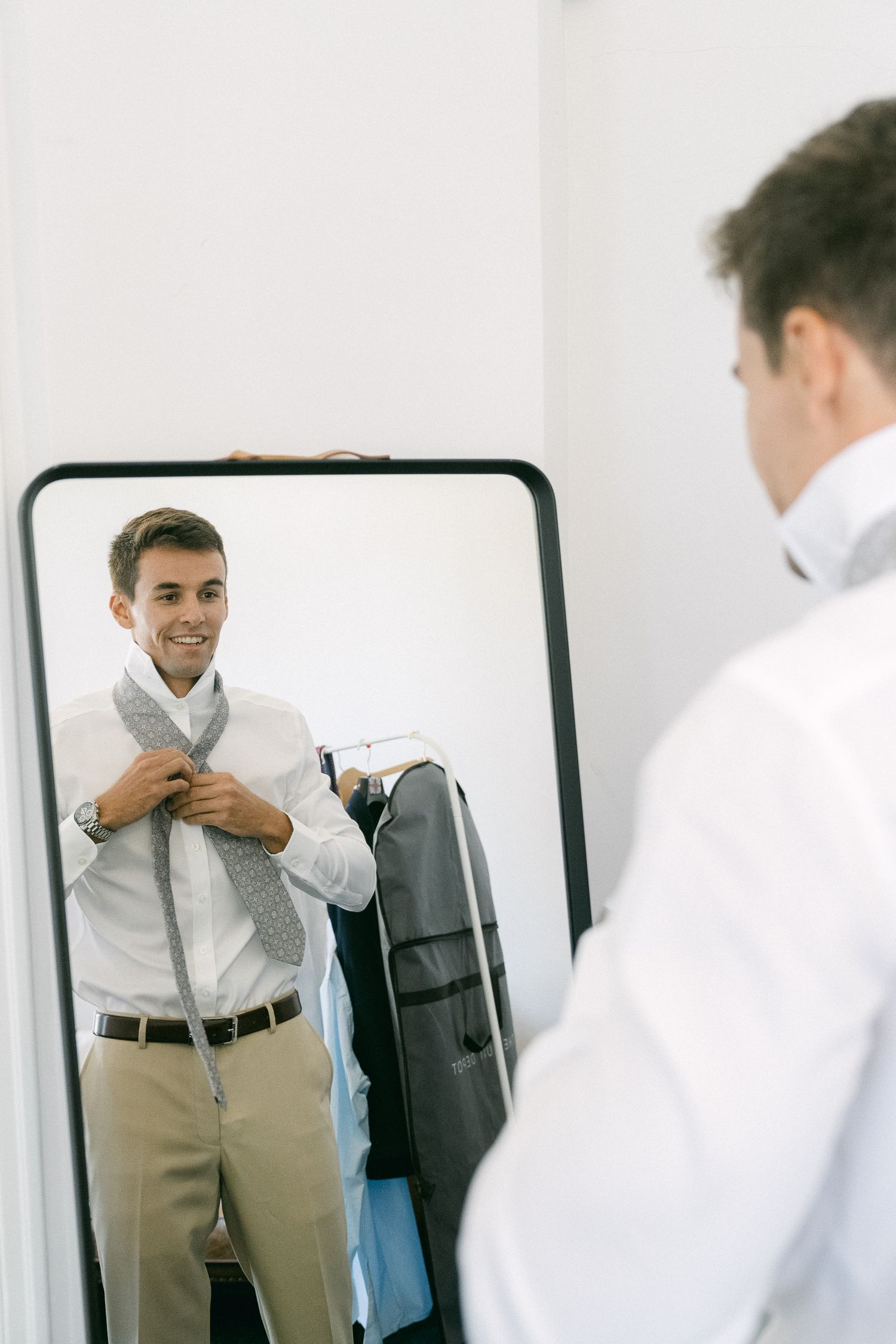 Young man smiling while tying a gray tie in front of a mirror, wearing a white dress shirt and khaki pants.