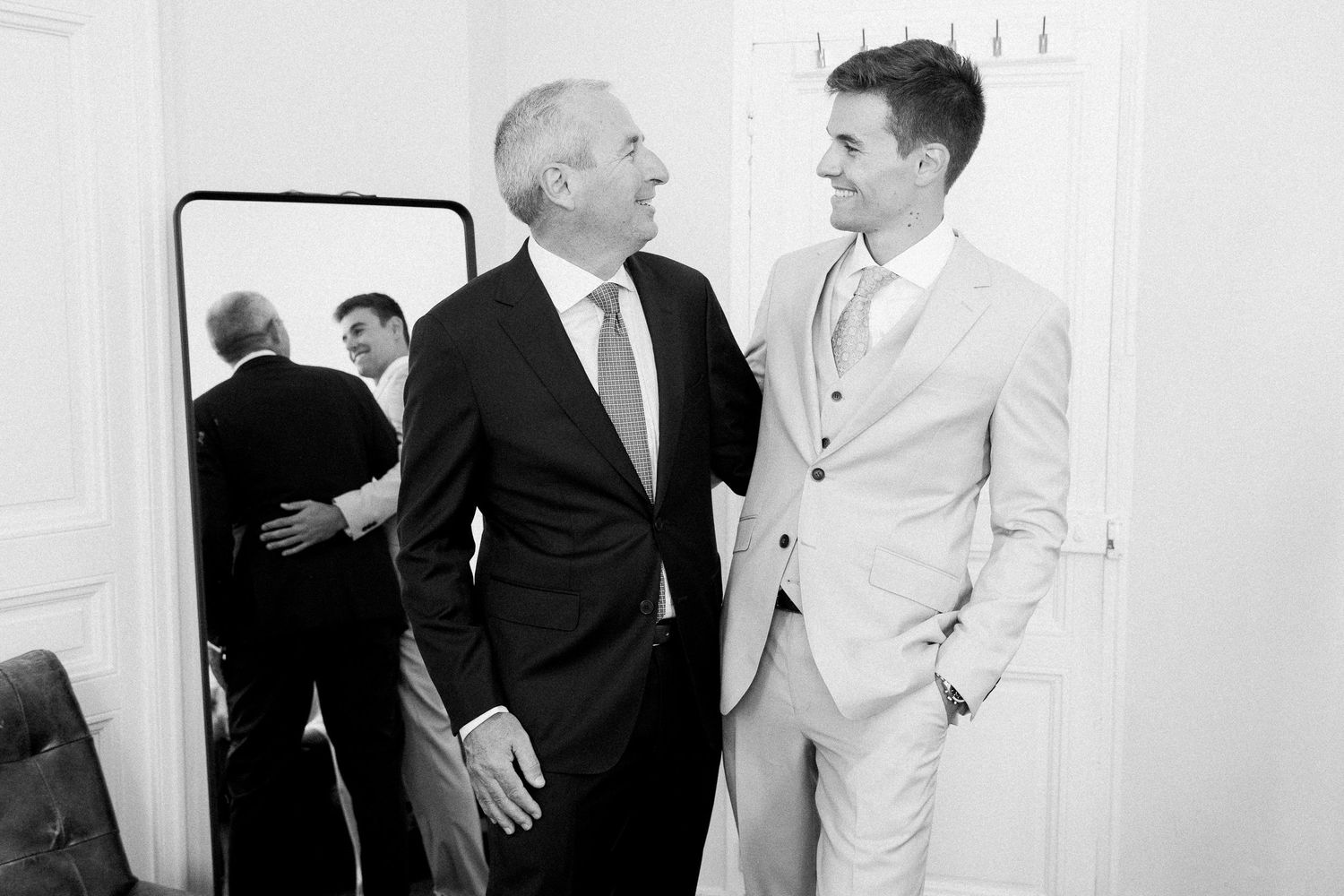 Two men in formal suits share a joyful moment beside a mirror in a black and white wedding photo.