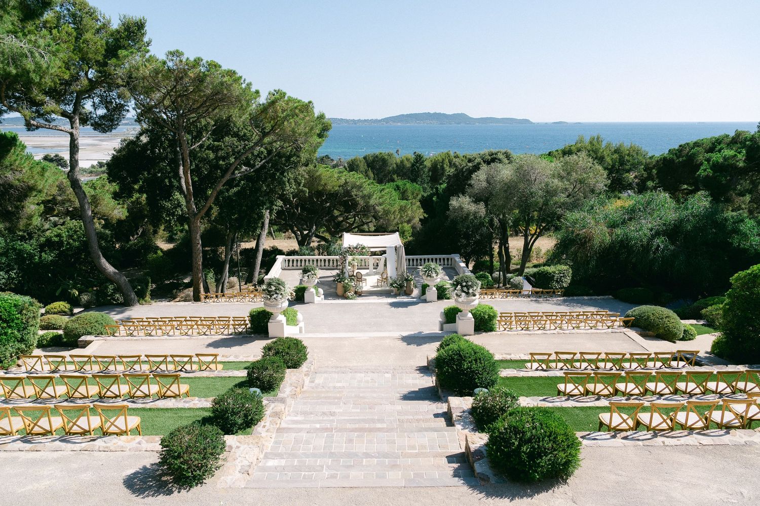Elegant outdoor wedding ceremony setup with manicured hedges, stone steps, and sea views in a Mediterranean garden.