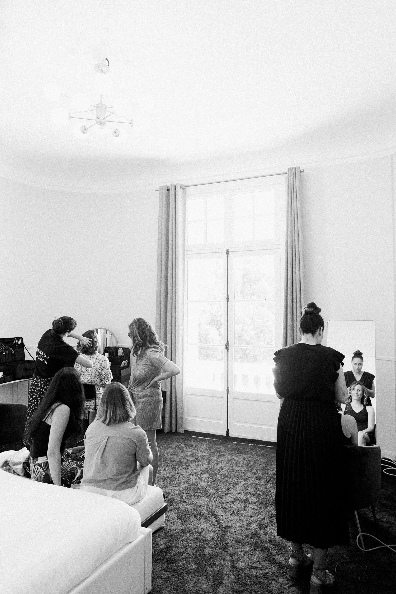 Black and white photo of women gathered in elegant room, one getting hair done near bright window with mirror.
