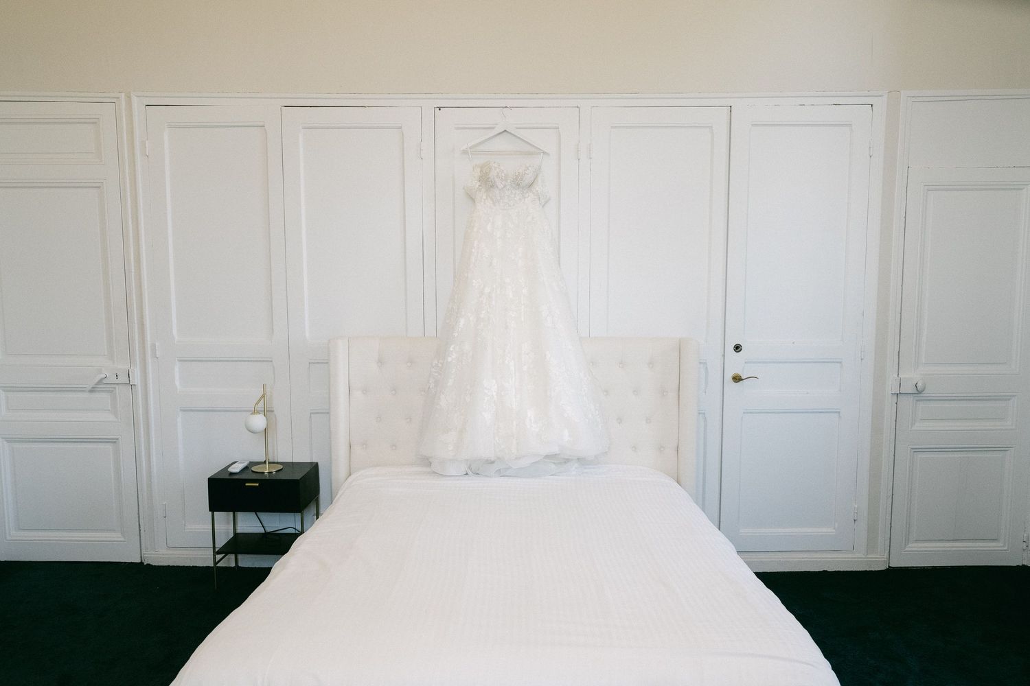 A white wedding dress hanging above a bed in a bright, elegant room with white paneled walls and doors.