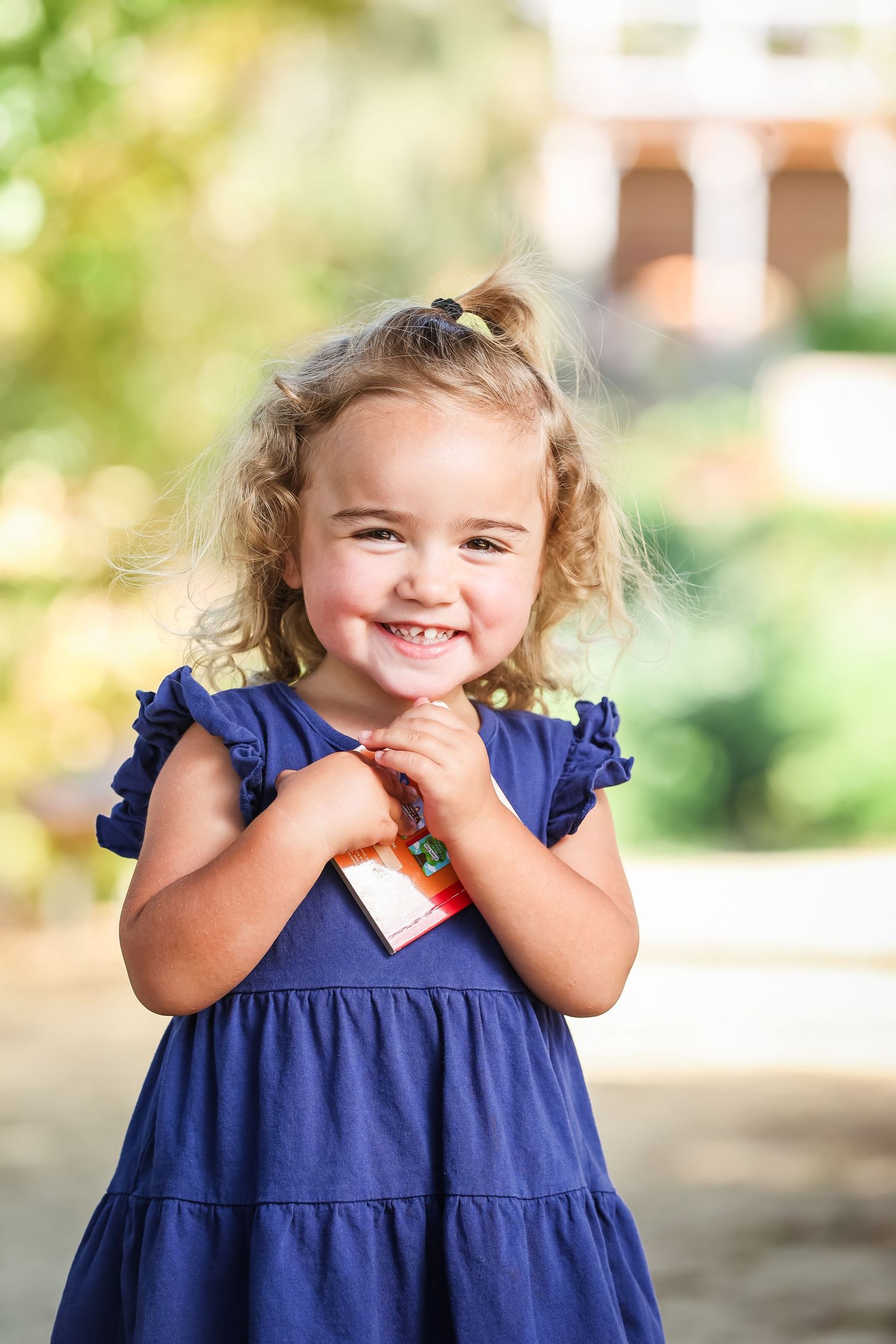 Smiling toddler girl with curly blonde hair wearing a blue dress, posing outdoors in a sunny garden setting.