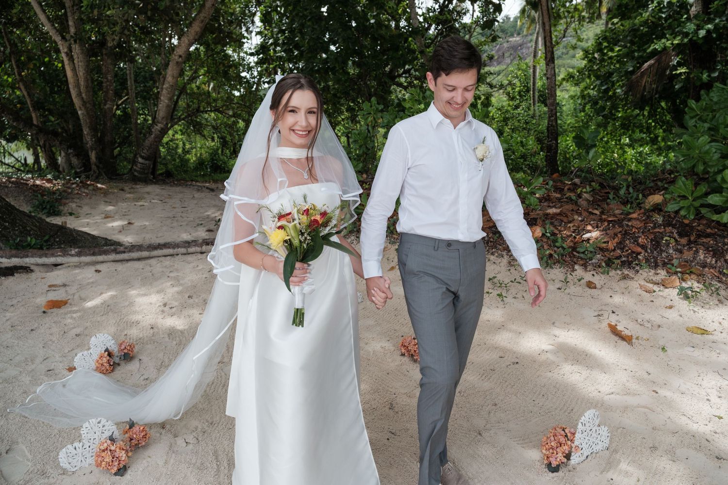 Bride and groom walking hand in hand on sandy beach path surrounded by tropical trees in wedding attire.