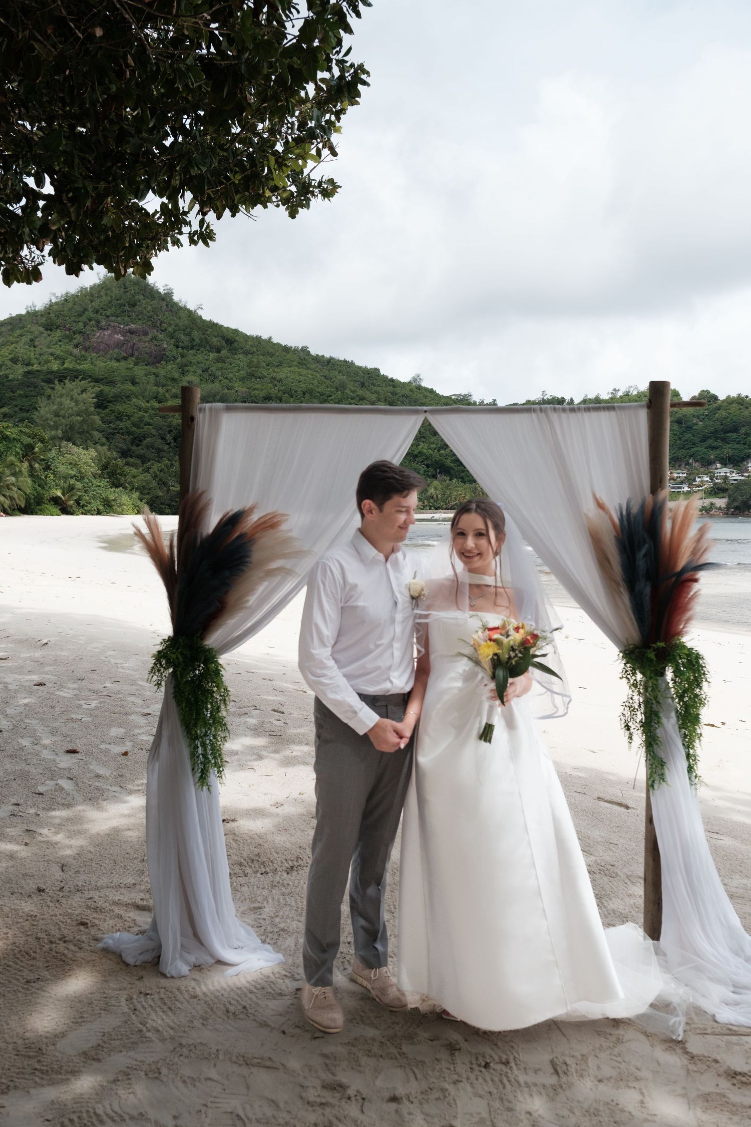 Bride and groom stand under a decorated beach wedding arch with white draping and tropical greenery.