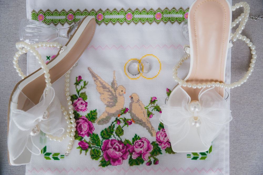 White bridal heels with pearl accents and bow beside embroidered cloth featuring doves, roses, and wedding rings.