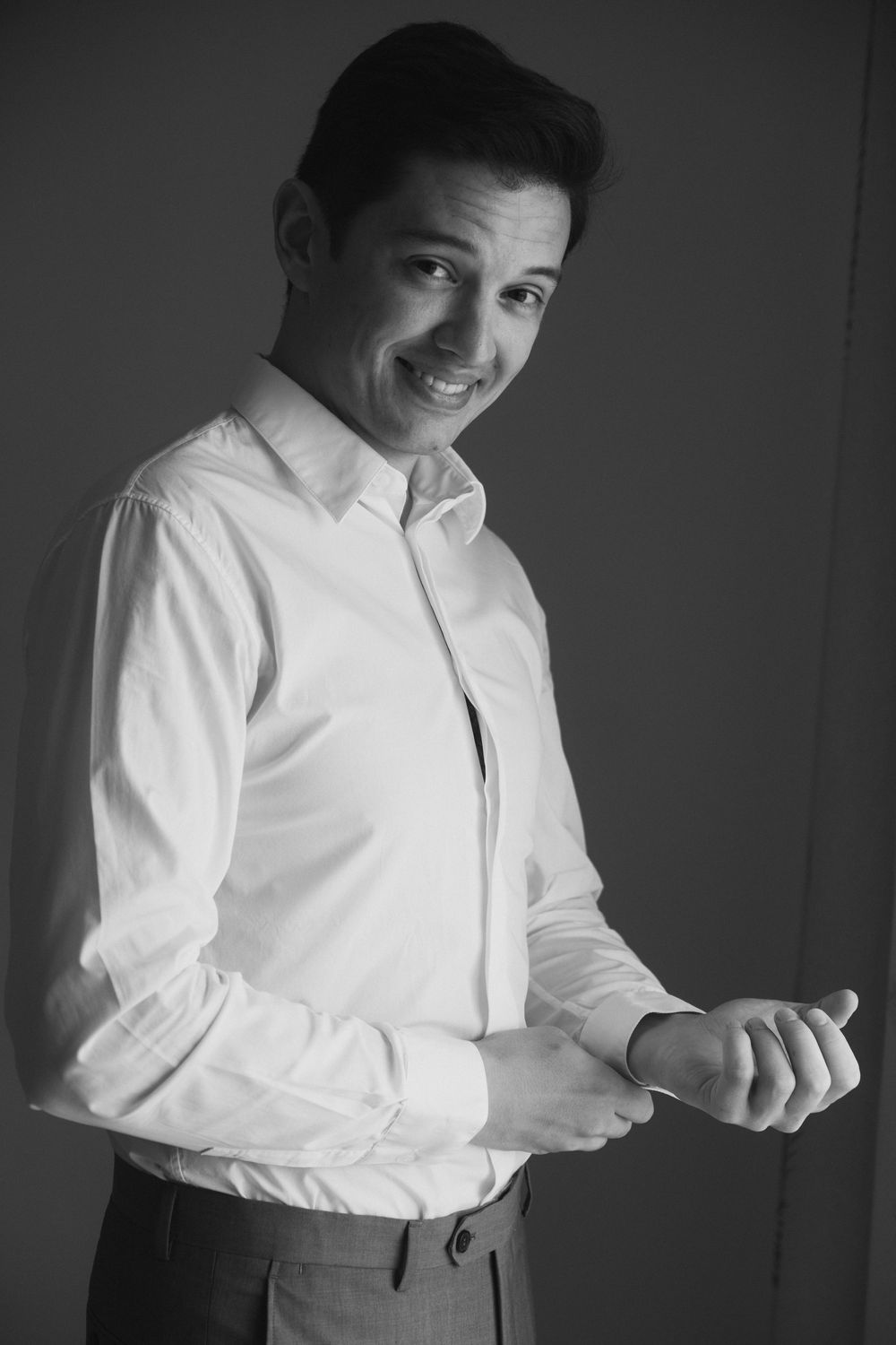 Black and white portrait of a smiling young man in a white dress shirt adjusting his cufflinks.