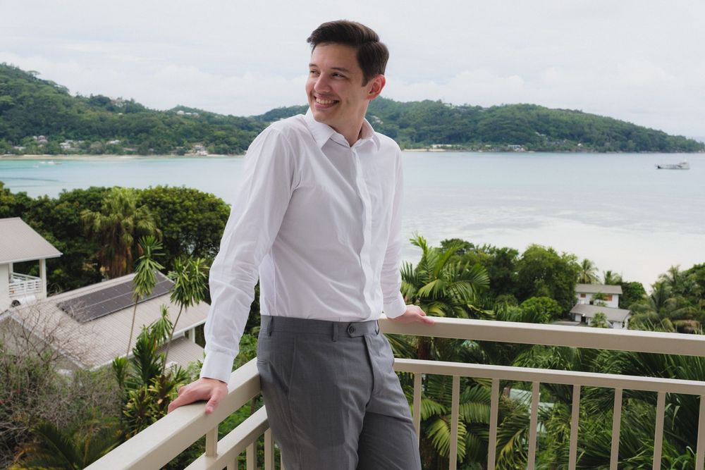 Young man in white shirt and gray pants stands on a balcony overlooking a tropical beach and lush greenery.