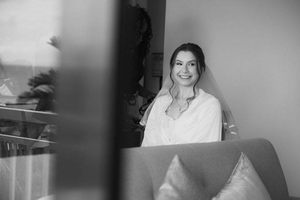 Black and white photo of a smiling bride in a white robe wearing a veil, seated in a bright room.