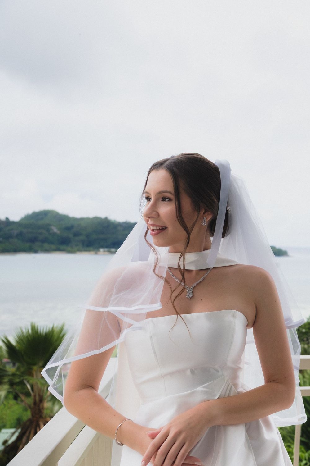 Smiling bride in white strapless gown and veil stands on balcony with ocean and tropical island backdrop.