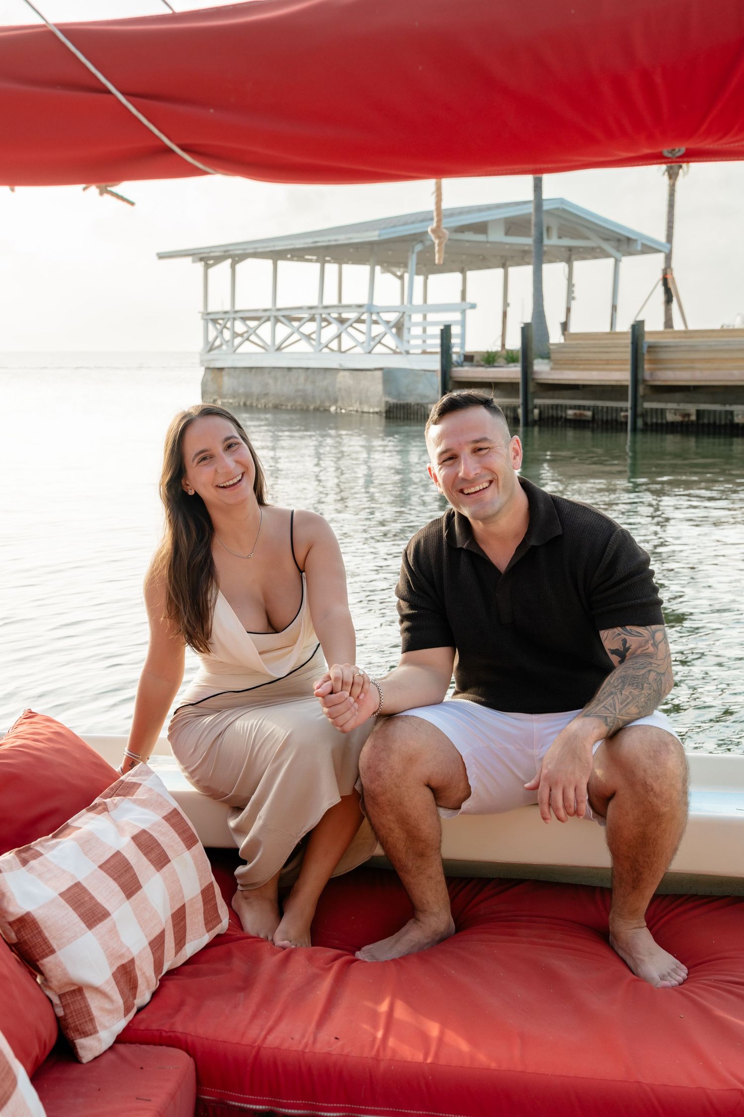 Smiling couple holding hands on a red boat lounge with a waterfront dock in the background.
