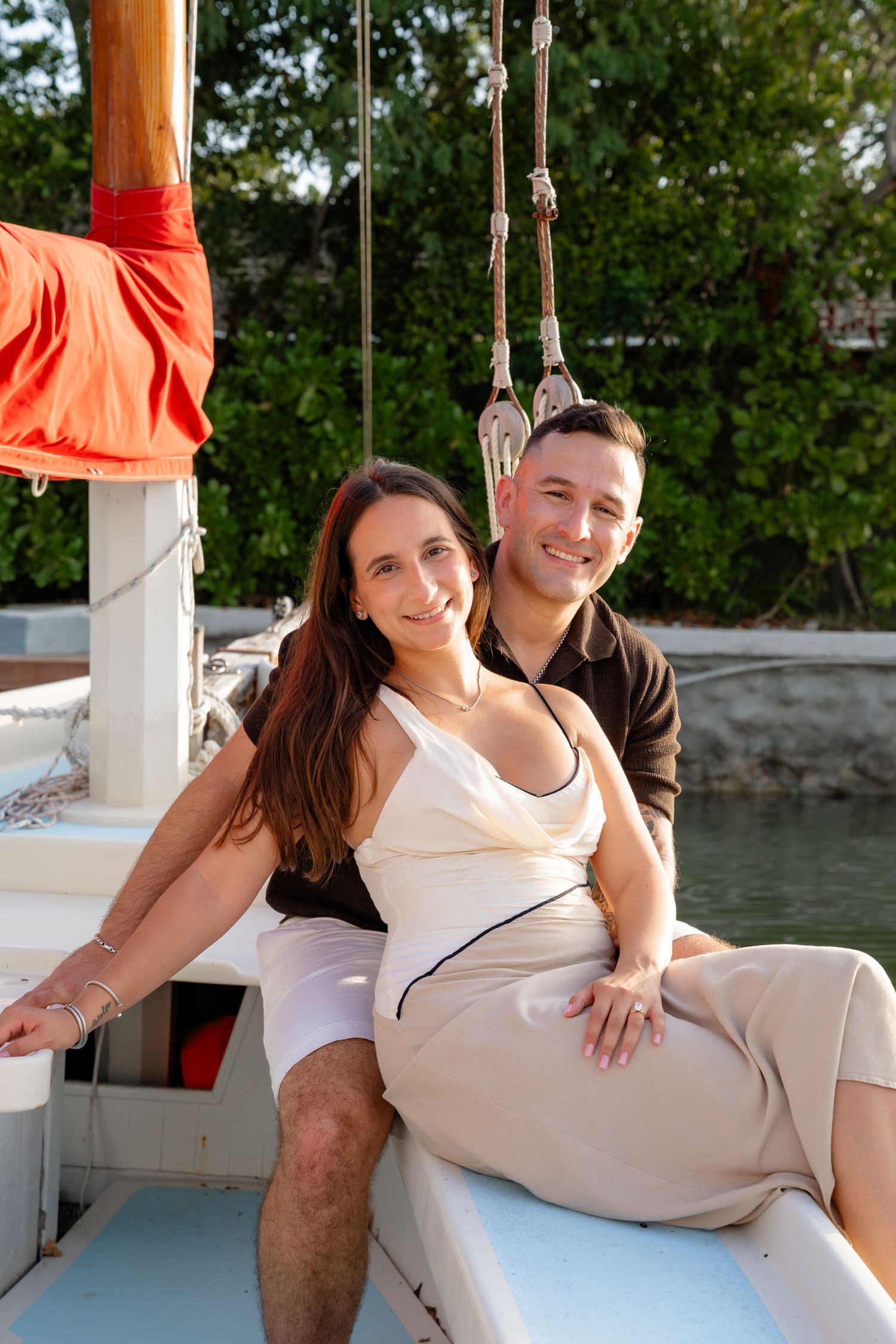 A smiling couple sitting together on a sailboat deck with green trees in the background on a sunny day.