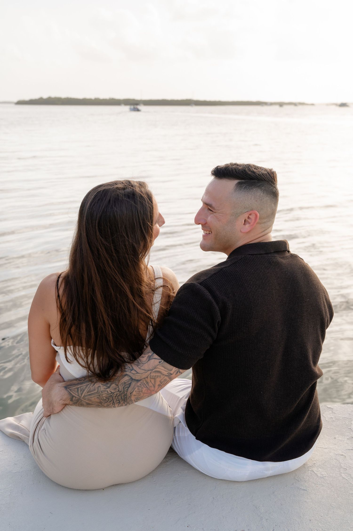 Couple sitting together on a white sand beach, smiling at each other with calm ocean water in the background.