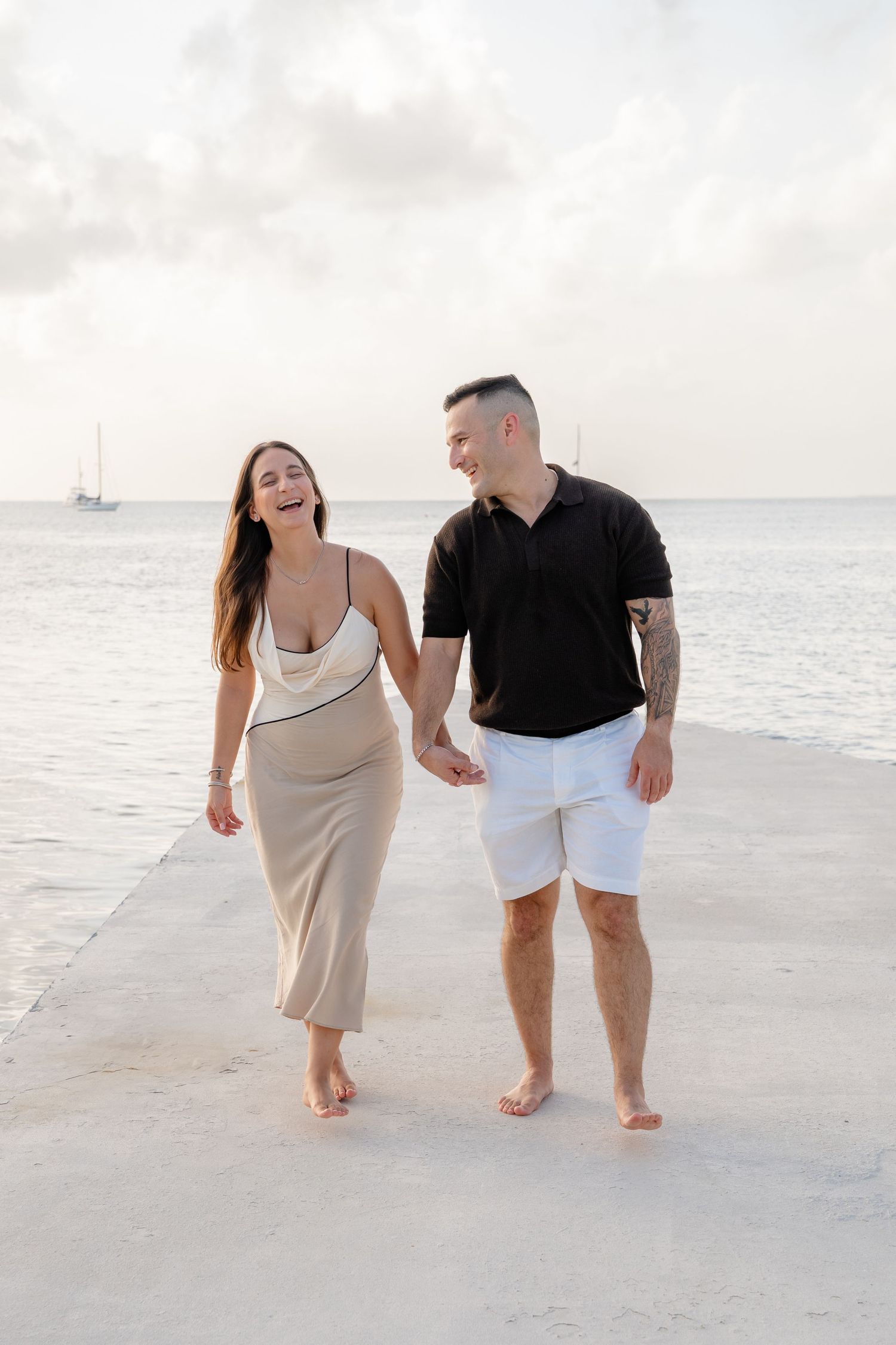 Happy couple holding hands and walking barefoot on a seaside pier, woman in beige dress, man in black polo and white shorts.