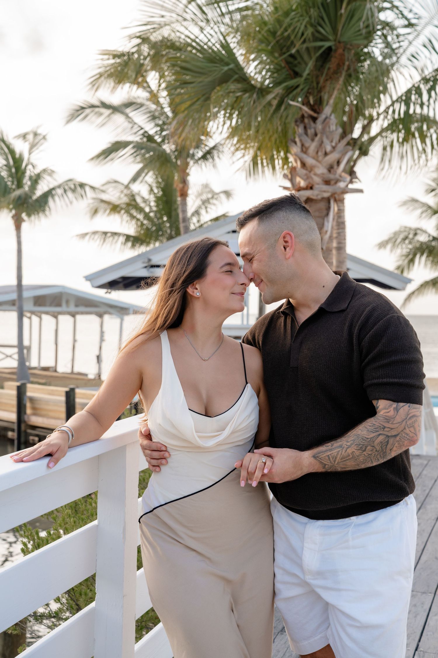 Couple sharing a romantic moment on a white railing deck with palm trees and ocean in the background.