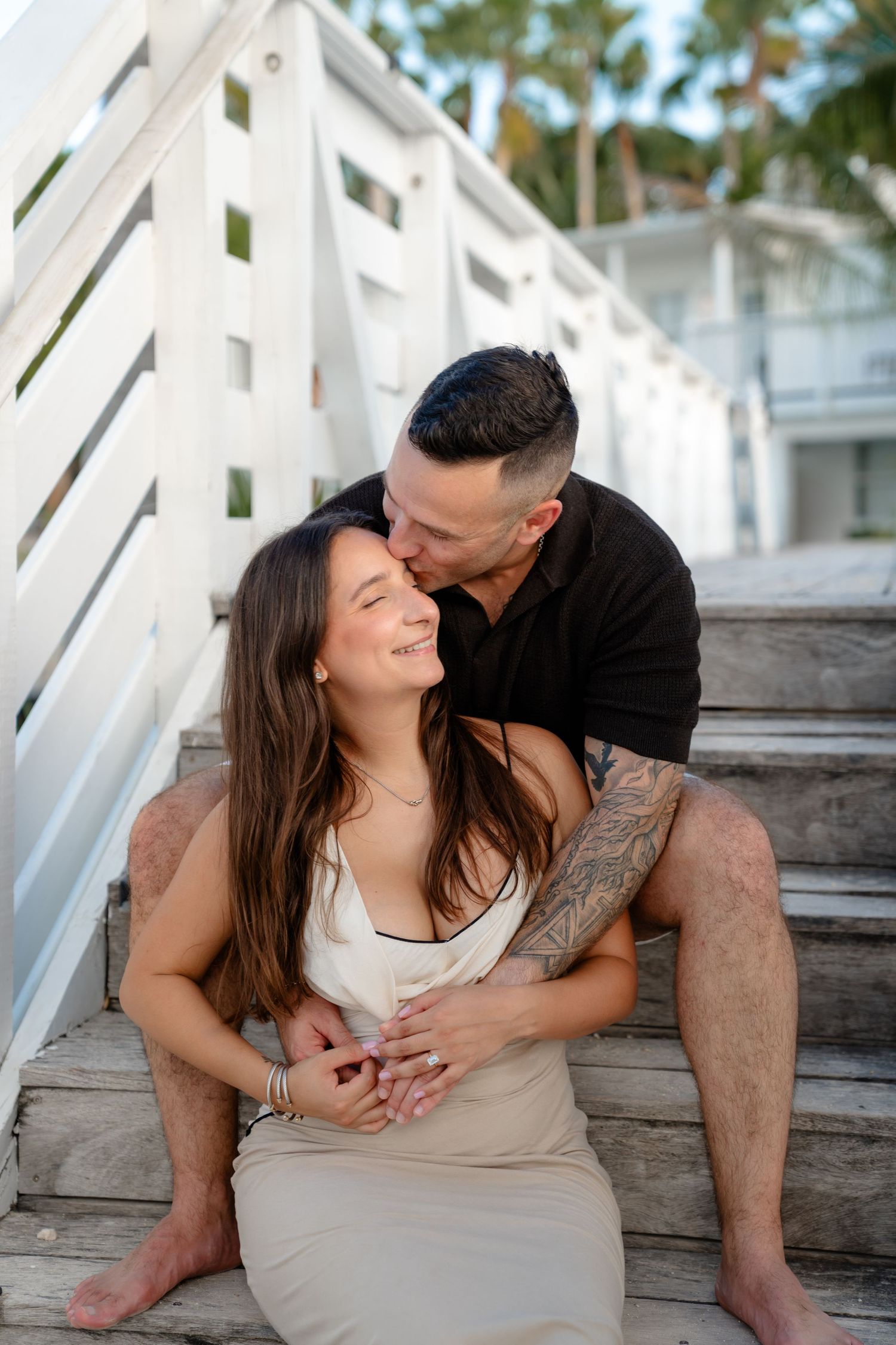 A couple sits on wooden steps, man in black shirt kisses woman's head while embracing her in a beige dress outdoors.