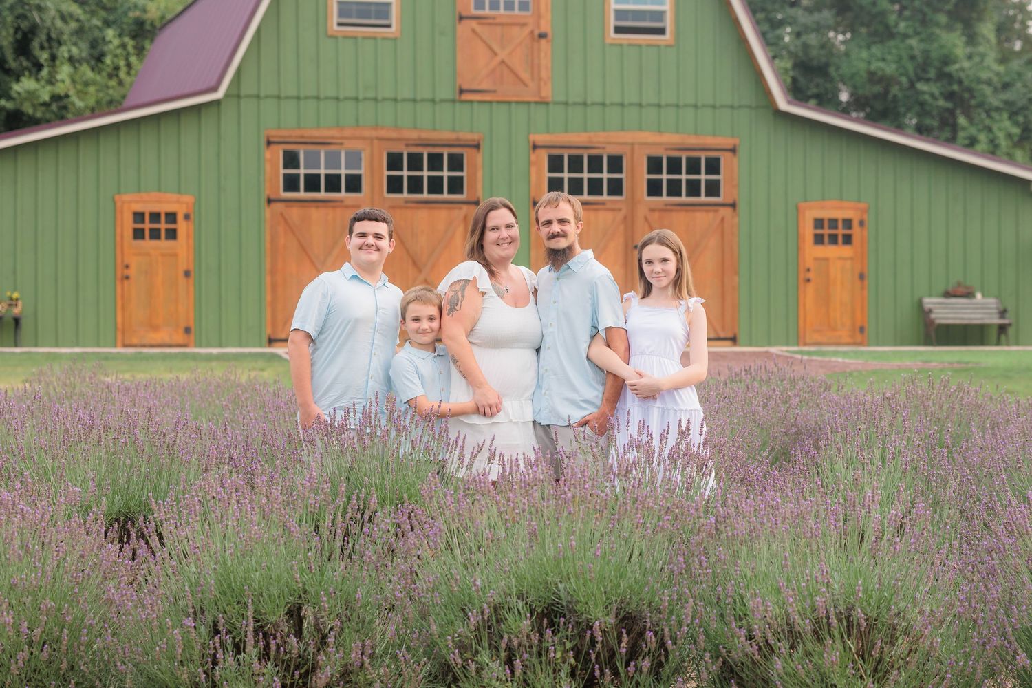 Family of five posing in lavender field before green barn, wearing coordinated white and blue outfits for outdoor portrait.