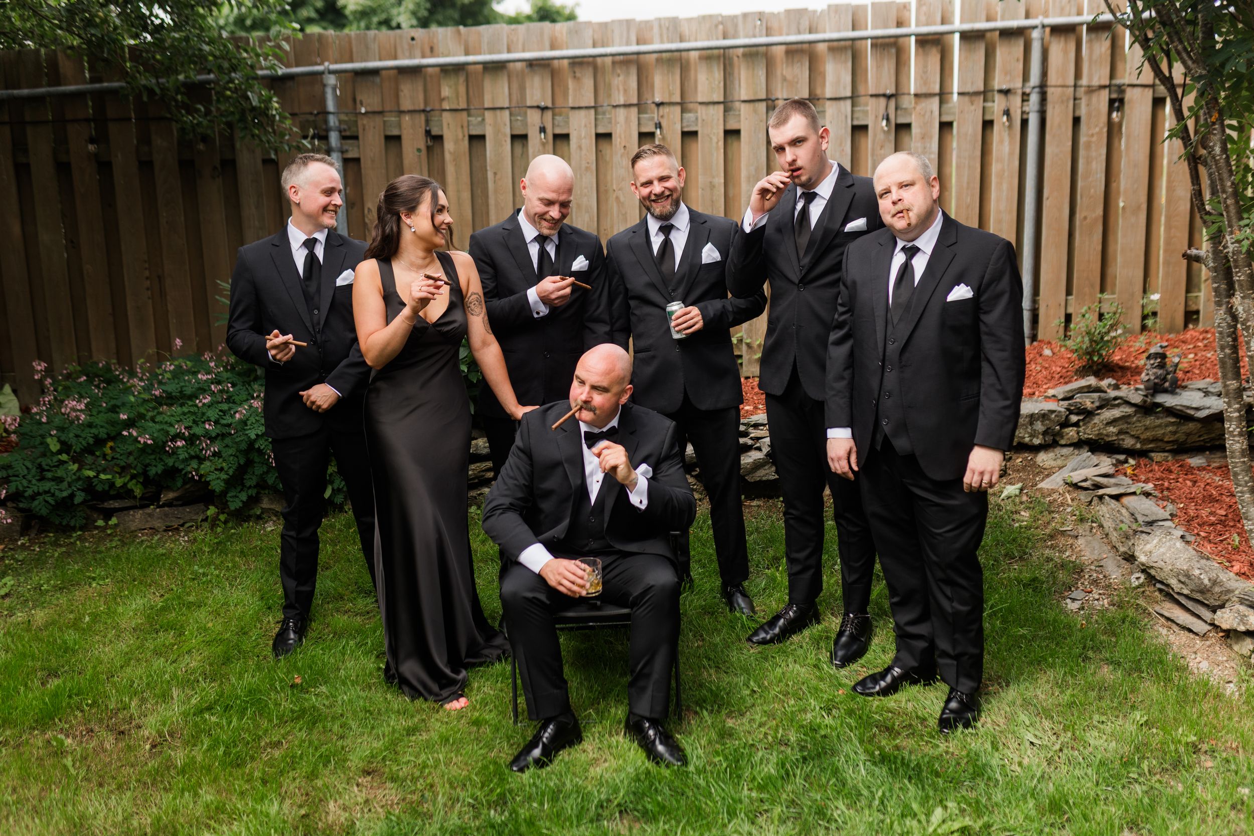 Wedding party in black formal attire posing outdoors in a backyard, with groomsmen and a bridesmaid smiling.