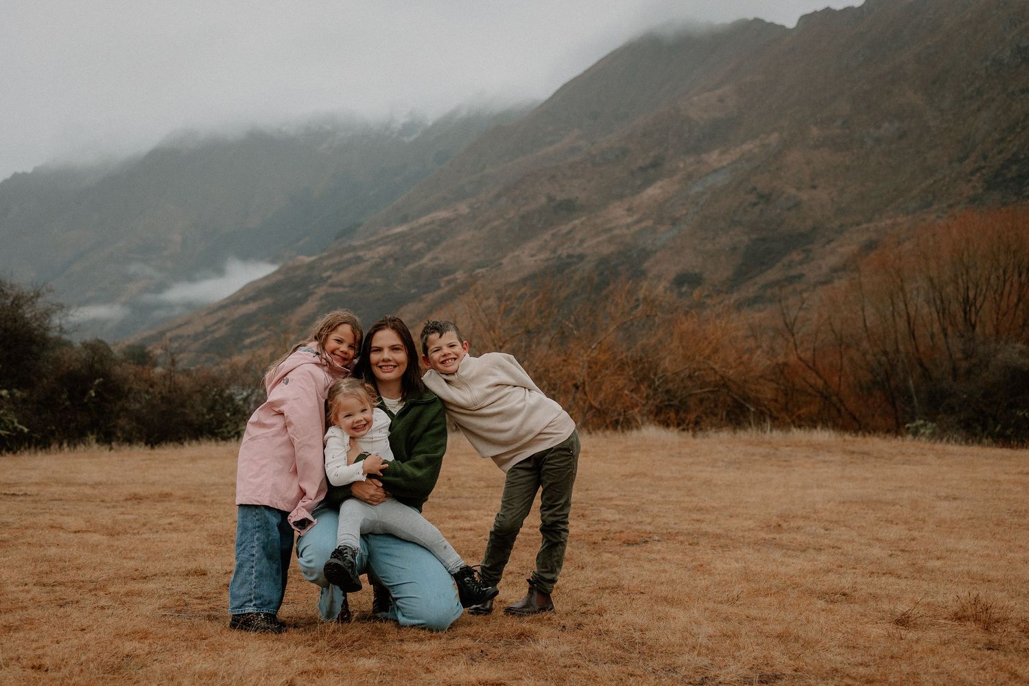 Family of four posing together in a misty mountain meadow during autumn, smiling warmly in casual outdoor attire.