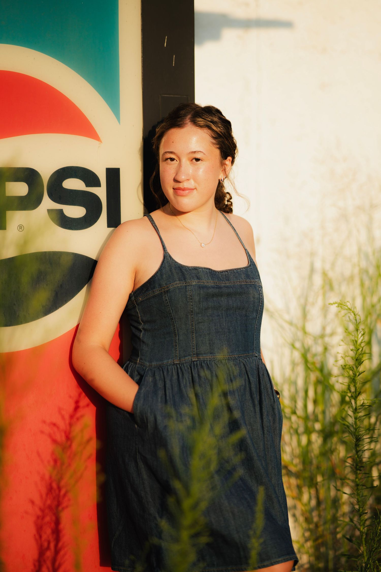 Young woman in denim sundress posing in front of a Pepsi sign outdoors in warm golden light.