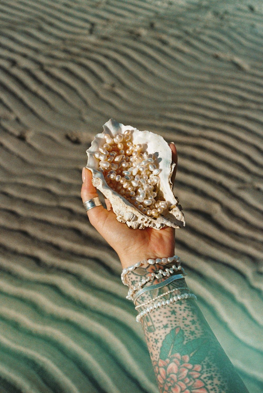 A hand adorned with bracelets holds a large seashell filled with pearls on a rippled sandy beach.