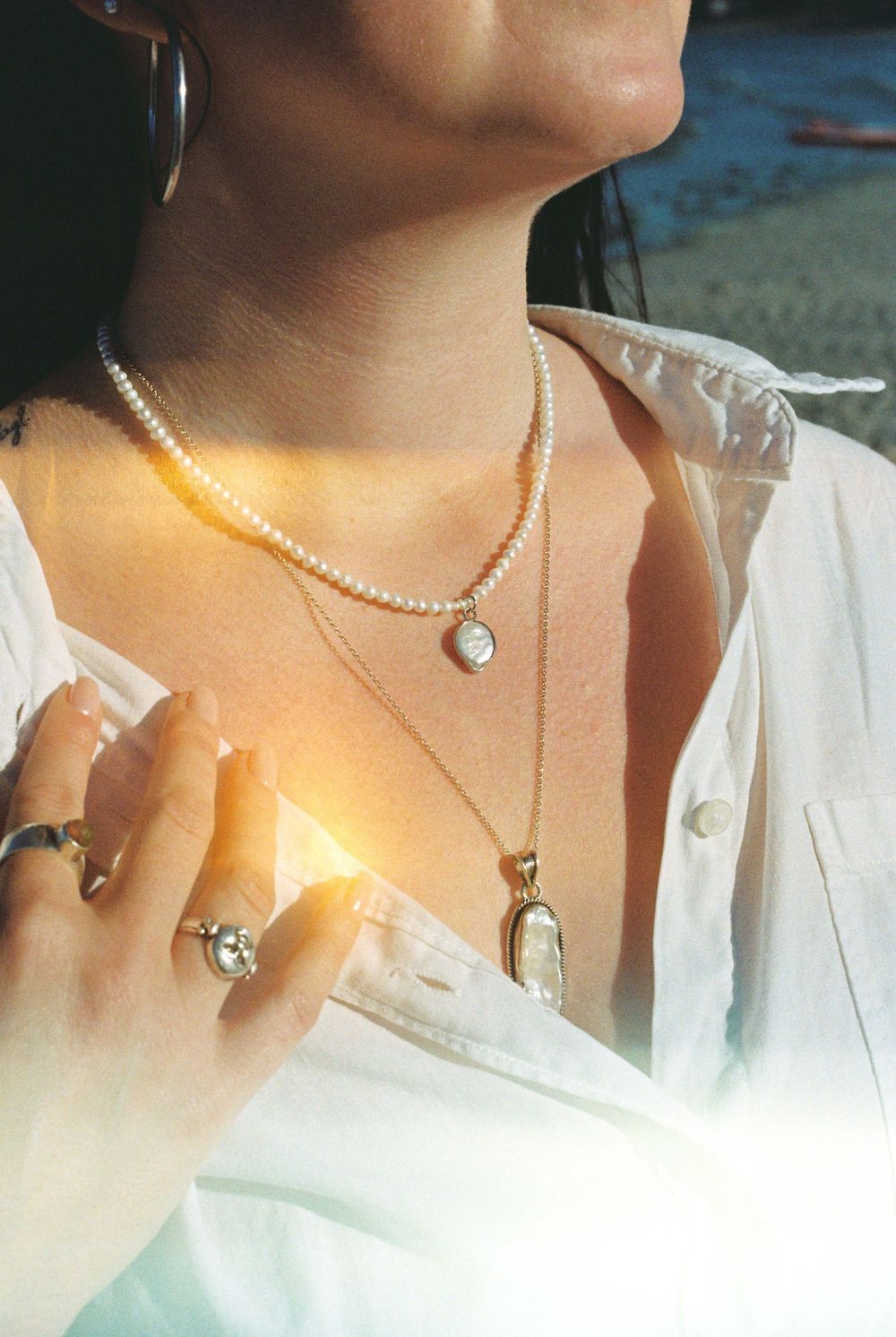 Close-up of layered pearl and gold necklaces with rings on woman wearing white shirt outdoors.