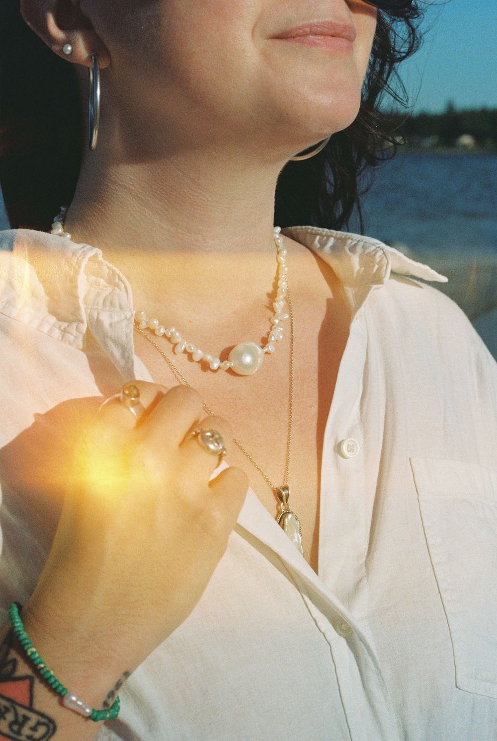 Woman wearing pearl beaded necklace and gold jewelry layers with hoop earrings near waterfront.