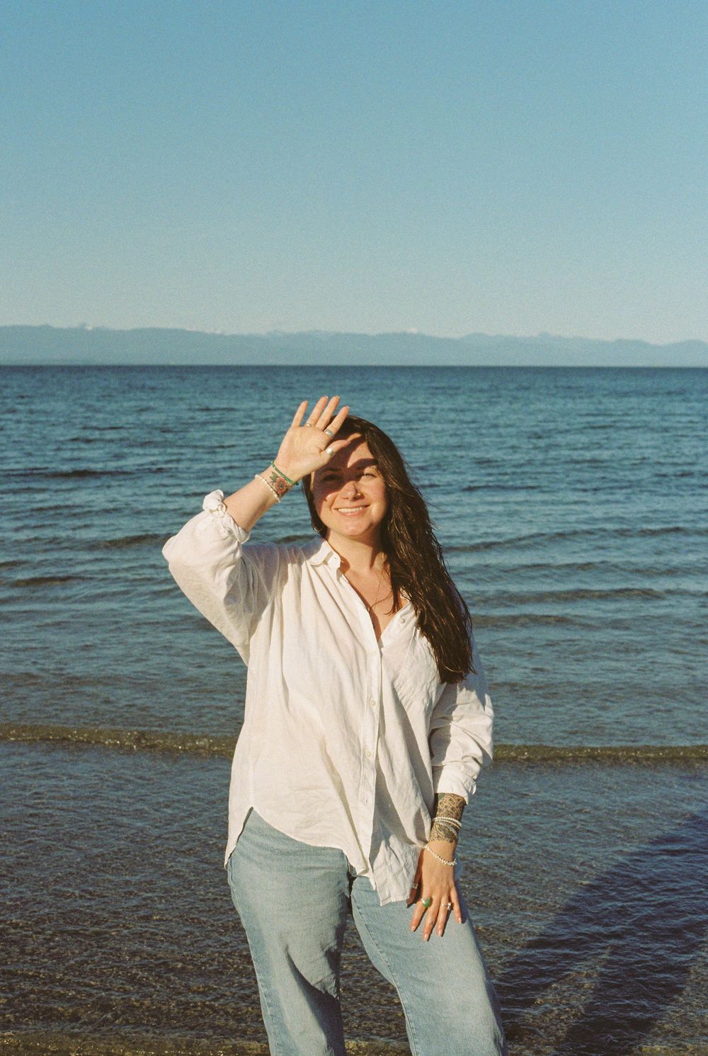 Woman in white shirt and jeans shielding eyes from sun while standing on a beach with blue ocean backdrop.