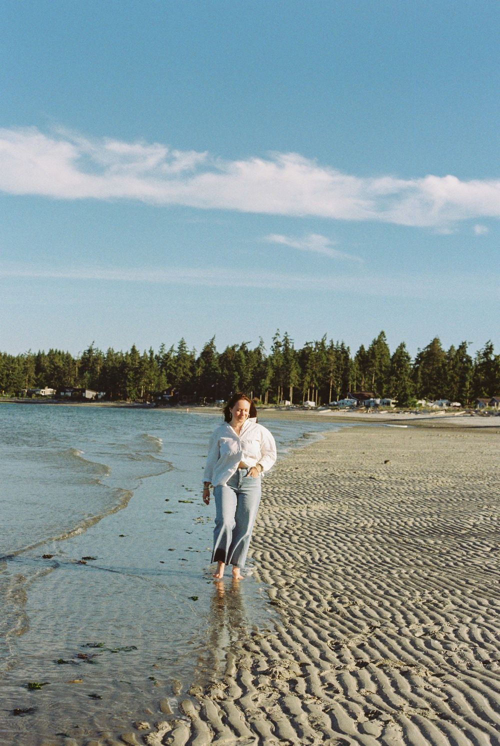 Woman in jeans and white shirt walking barefoot on a sunny sandy beach with trees in background.