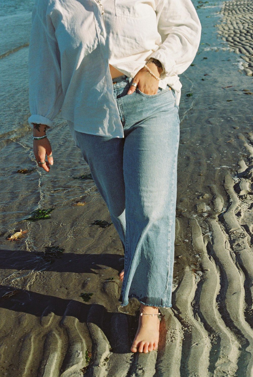 Close-up of a woman in light wash jeans and white shirt walking barefoot on a sandy beach.