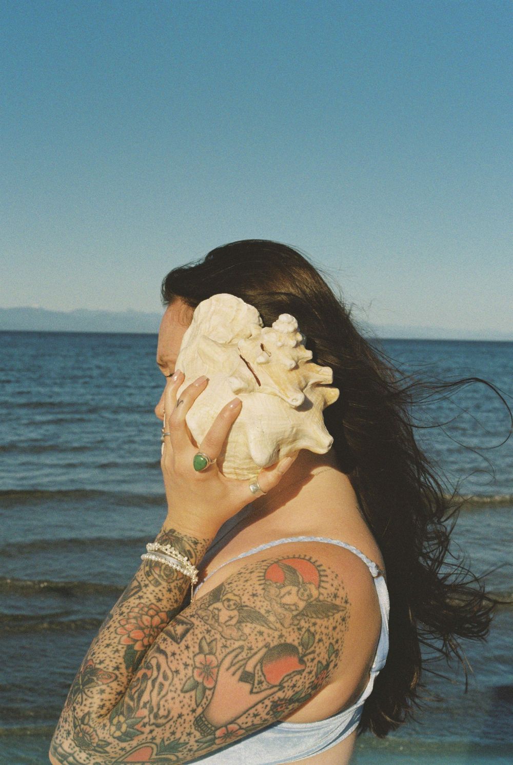Tattooed woman holding a large conch shell to her ear on a sunny beach with blue ocean water in the background.