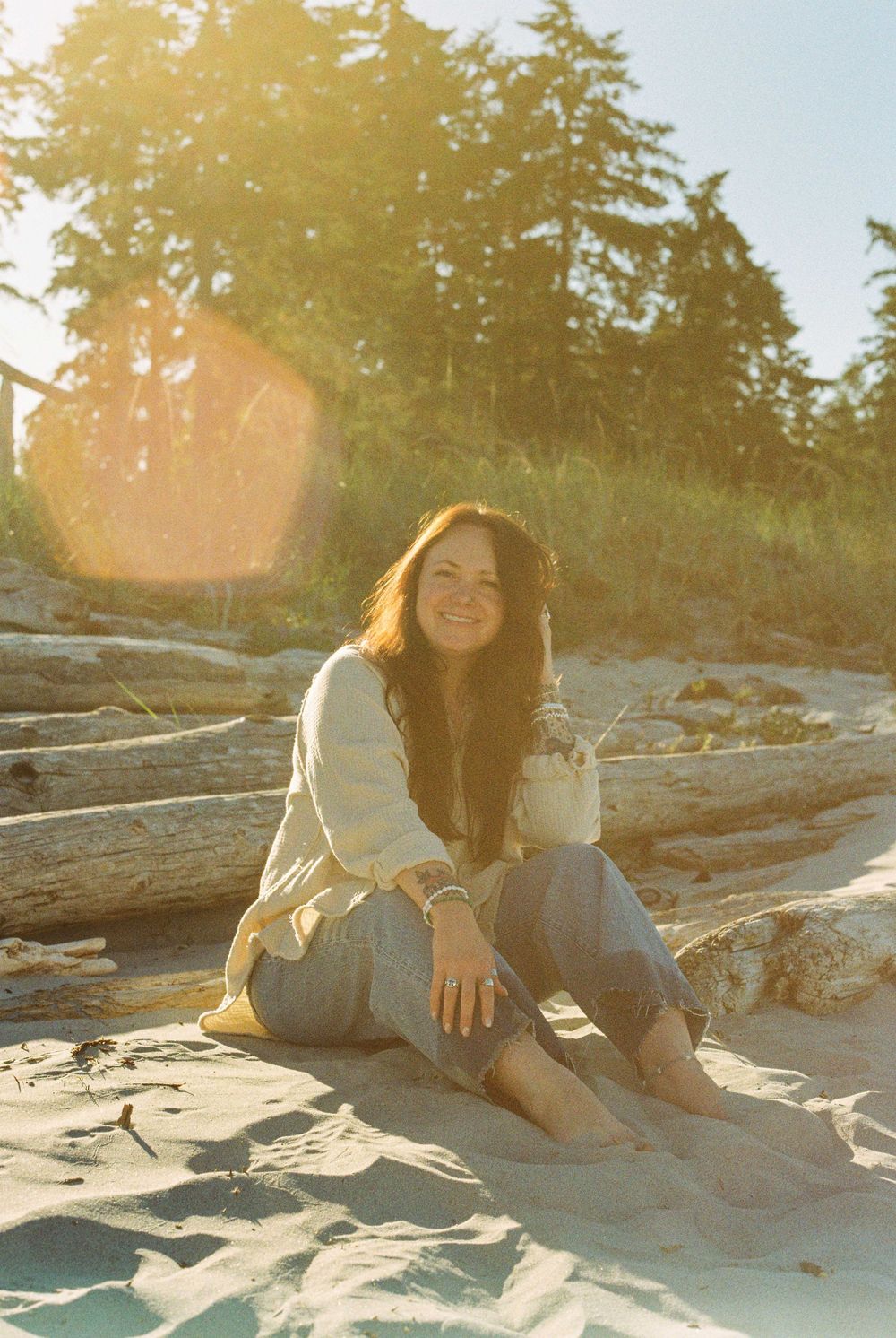 Woman with long brown hair sitting on sandy beach at golden hour, smiling, wearing linen outfit with driftwood and trees behind.